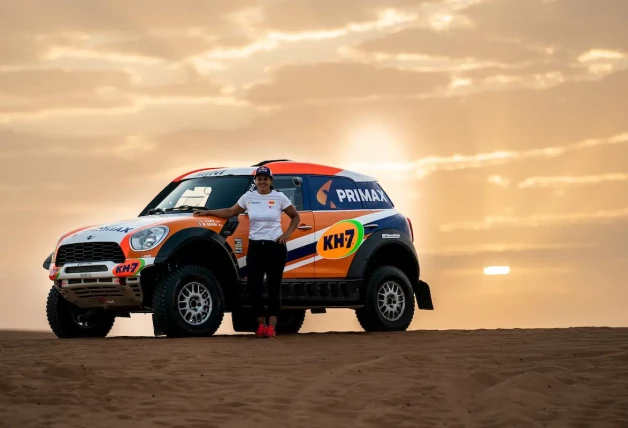 Imagen de la piloto Laia Sanz con ropa deportiva, de pie al lado de un vehículo de rally todoterreno en un entorno desértico.