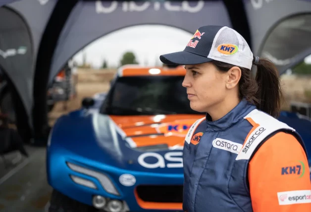 La imagen muestra a la piloto Laia Sanz de perfil, enfocada y seria, con una gorra y una chaqueta de carreras, en un entorno de competición de motor con un coche de rally azul y naranja desenfocado en el fondo.