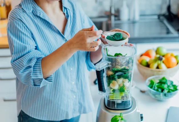 Mujer que agrega polvo verde espirulina durante la preparación del batido verde en la cocina