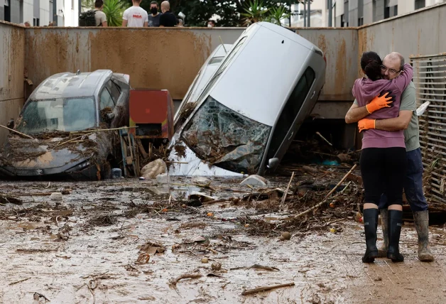 Un hombre y una mujer se abrazan junto a un montón de coches destrozados por la DANA.