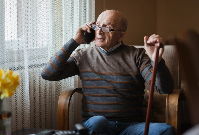 Anciano con bastón sentado en una butaca mirando por la ventana y hablando por teléfono.
