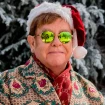 Elton John en un decorado navideño vistiendo abrigo y gafas de sol.