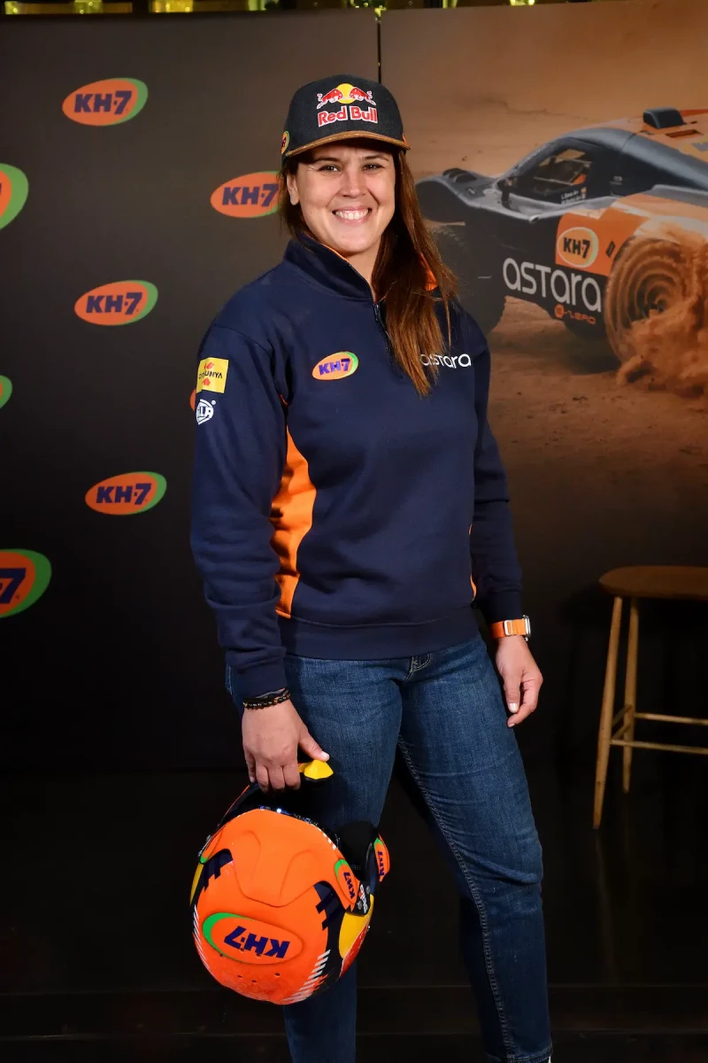 La imagen muestra a la piloto Laia Sanz de perfil, enfocada y seria, con una gorra y una chaqueta de carreras, en un entorno de competición de motor con un coche de rally azul y naranja desenfocado en el fondo.