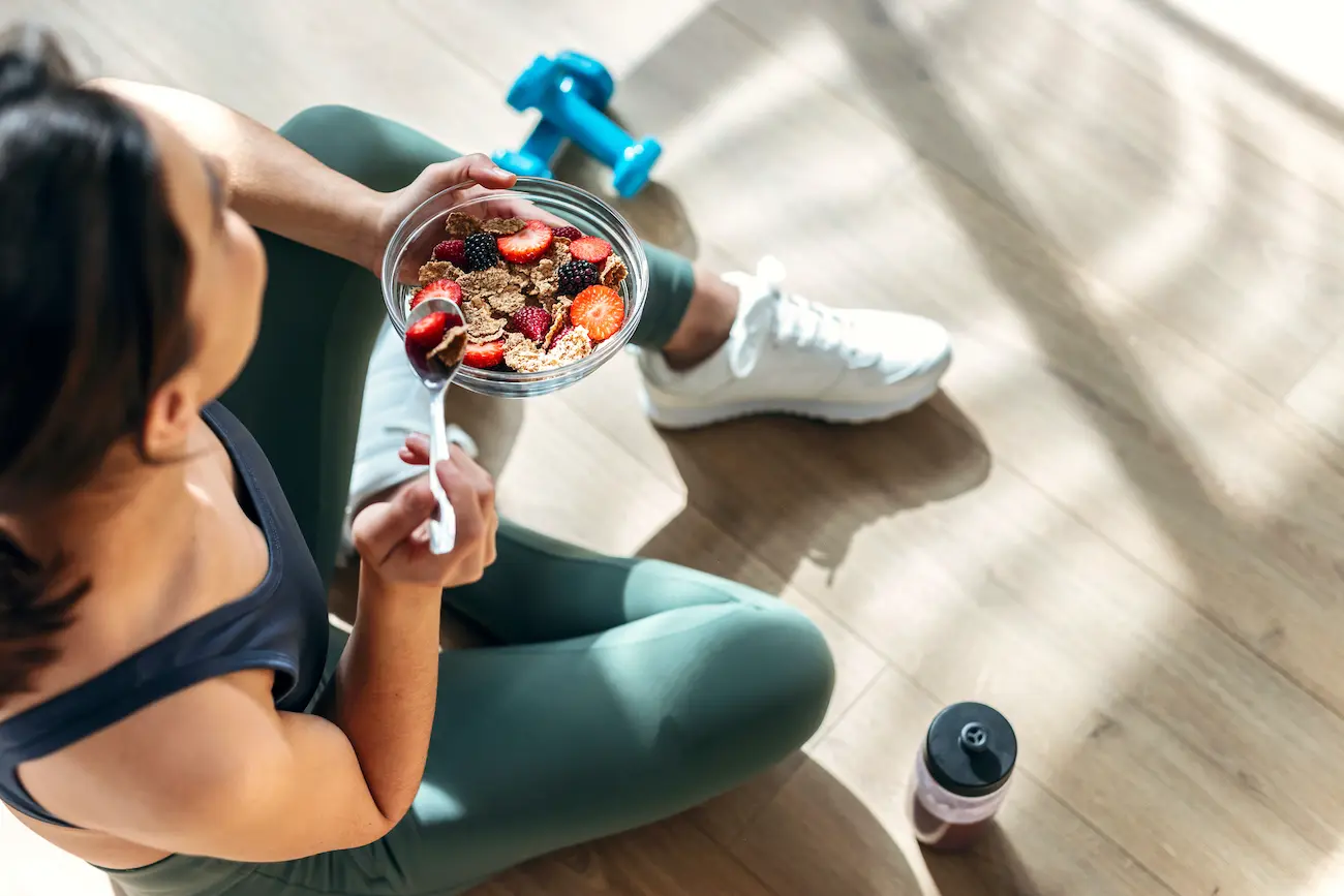 Mujer atlética comiendo un tazón saludable de muesli con fruta sentada en el suelo en la cocina de su casa